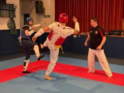 Adults practicing taekwondo kicks and drills in a busy indoor training hall, with their coach supporting