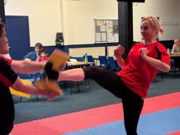 A woman is practising a high kick, she has kicked through a wooden plant held by the coach